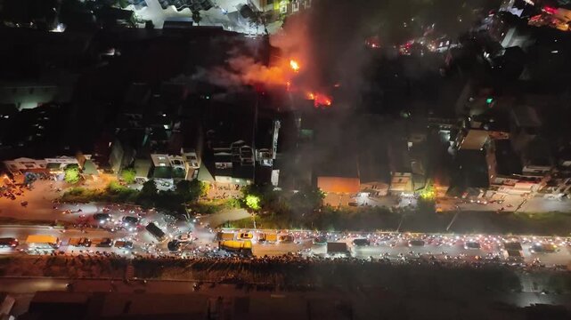 Aerial night view of congested Hanoi street beside active fire scene, dense traffic illuminated by headlights and emergency glow.