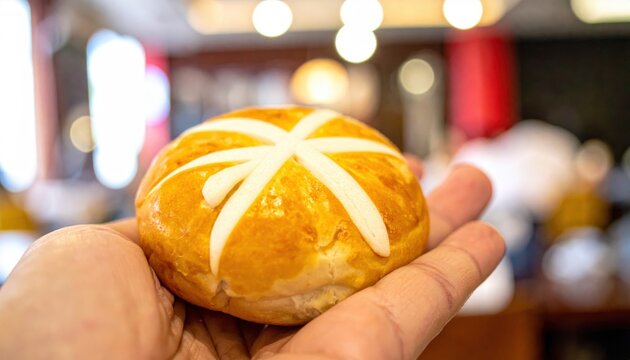 A hand holds a perfectly formed char siu bao, a delicious steamed bun filled with barbecue pork, presented with a golden crust and white icing cross.