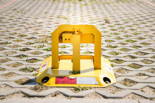 Yellow parking boot secured on a textured pavement with grass growing between hexagonal concrete blocks under bright sunlight in an outdoor setting