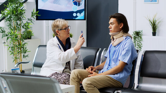 Senior doctor examining asian patient, checking eye vision during checkup visit consultation in hospital waiting room. Injured patient wearing cervical neck collar reciving medicine support