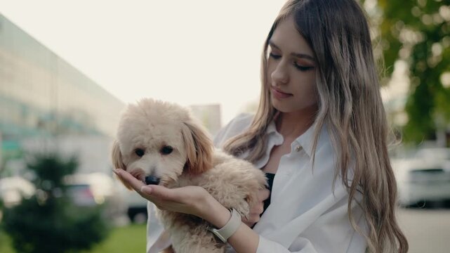 A young woman gently holds her small, fluffy dog in a park. She lovingly offers a treat from her hand, creating a heartwarming scene of connection and care. The dog enjoys the treat