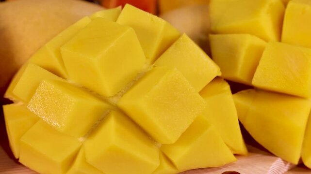 Fresh mango cubes on wooden board with pomegranate seeds and summer fruits in background.