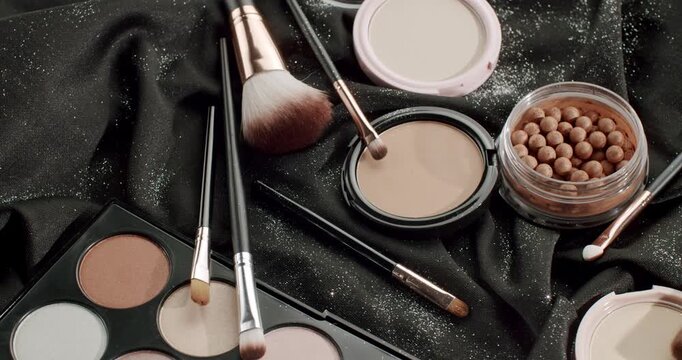Variety of cosmetic products and makeup brushes rotating in slow motion, including eyeshadow palettes, face powder, and bronzer pearls on a luxurious black sparkling fabric background