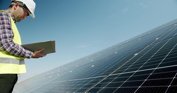 Male engineer inspecting solar panels. Positive adult man in uniform checking photovoltaic panels and making notes in journal during work on solar power station