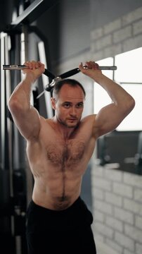Strong athletic man with a bare torso performing a pulldown exercise on a cable machine, intensively training his arm and back muscles during a strength workout in a modern fitness club, vertical