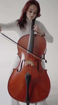 A beautiful woman sitting and playing the cello in a studio on a white background. Closeup view of violoncello and hands of musician, A female cellist plays the cello. Vertical video.