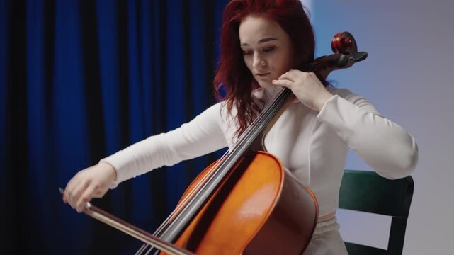 A beautiful woman sitting and playing the cello in a studio on a black blue background. Classical music playing stringed classical musical instruments, Female cellist practicing solo piece.