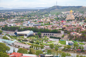 A panoramic view of Tbilisi, Georgia showing the golden-domed Holy Trinity Cathedral, riverside parks, modern Rike Park structures and historic neighborhoods set against green hills and city skyline © evannovostro