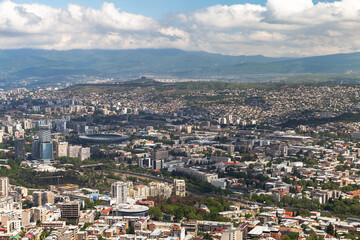 Fototapeta premium Expansive aerial view of Tbilisi city with a prominent stadium, mixed residential and commercial buildings, river corridor and distant green mountains under a partly cloudy sky. Georgia