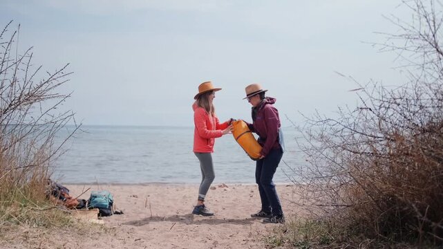 two women standing front view beach unpacking tent from backpack near lake, preparing campsite together, outdoor travel activity, hiking trip, teamwork, camping setup and exploration