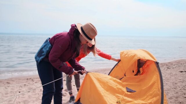 two women setting up tent close-up near lake shore, fixing poles and adjusting fabric, outdoor camping activity, hiking trip, teamwork, preparation and travel exploration in nature