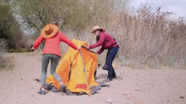 two women struggling setting up tent front view near lake, pulling fabric and adjusting poles, outdoor camping activity, teamwork challenge, minor conflict, problem solving and travel experience
