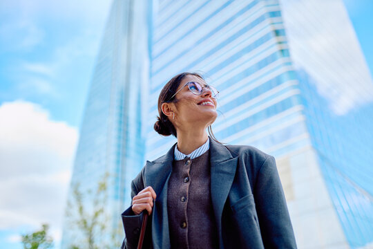 Happy businesswoman wearing glasses looking up, standing in front of office architecture. Confidence and success in business