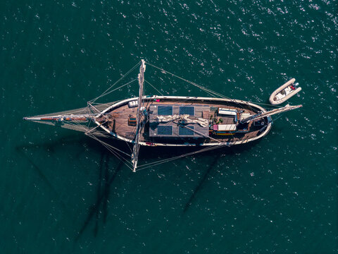 Aerial view of a large wooden sailing vessel with solar panels on deck and a small inflatable dinghy alongside on deep blue water.