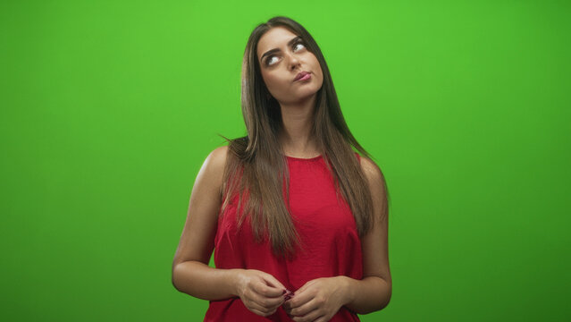 Young hispanic woman with hands clasping and slight head tilt, looking upward and fiddling fingers in a green screen studio; thoughtful reflection.