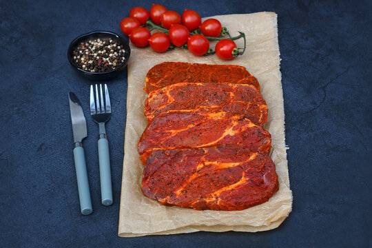 Freshly marinated pork neck steaks on baking paper with tomatoes, pepper, and cutlery. Preparation for grilling.