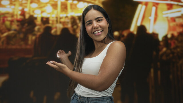 Young woman smiling with open hands presenting a nighttime street fair scene amid bright lights and crowded stalls; joyful welcome.