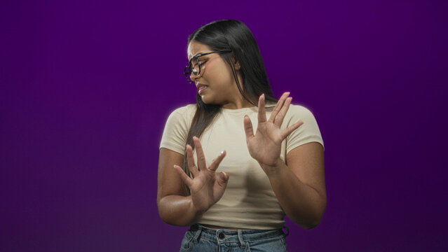 Young woman with glasses and hands raised, recoiling with a disgusted facial grimace, wearing beige tee and jeans in a purple studio; disgust avoidance.