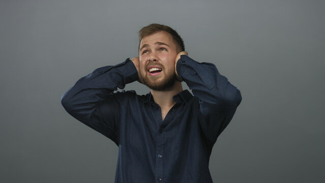 Young caucasian man pressing hands over ears and wincing in gray studio wearing navy shirt; pain noise sensitivity.