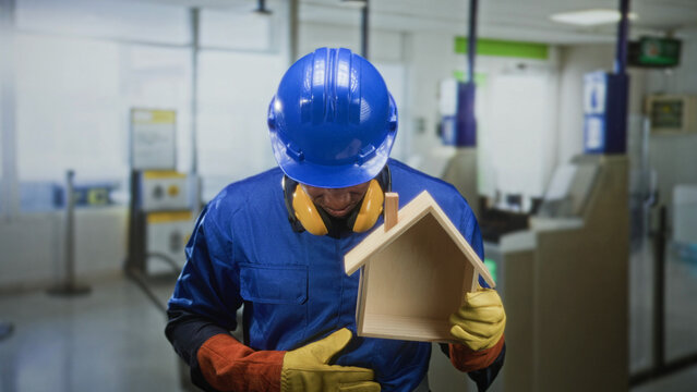 Black builder in blue hardhat and coveralls holds a small wooden house model while adjusting yellow ear protection and glove in airport terminal; home repair pride.