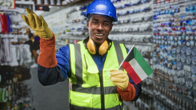 Man construction worker wearing hardhat and high visibility vest holds kuwait flag and waves gloved hand in building aisle; pride solidarity.