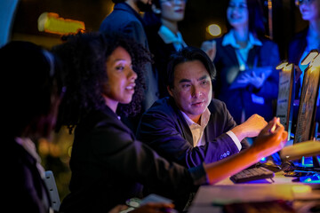 Fototapeta premium Dedicated male and female professionals discussing project strategy at night. Crowded IT agency workplace with diverse employees analyzing data on glowing screens.