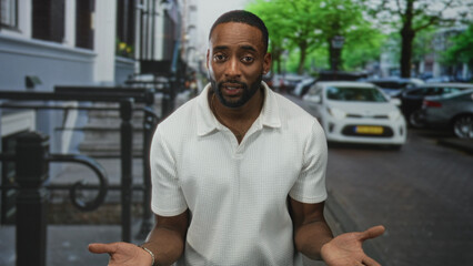 Obraz premium Man with hands clasped at waist wearing white polo and bracelet, standing on street sidewalk among parked cars and trees, looking at camera; thoughtful reflection.