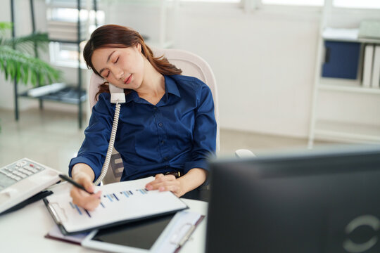 Weariness in Office: An office worker, eyes closed and resting, leans onto the phone, as she continues to work, showcasing the relentless demands of the modern office environment.