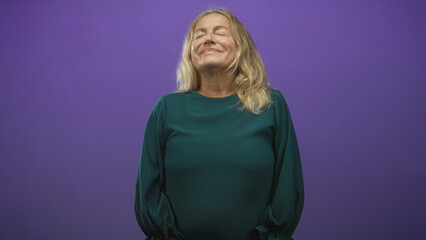 Fototapeta premium Senior woman with eyes closed and a slight smile, hands gently resting in a green blouse in a purple studio; serenity reflection.