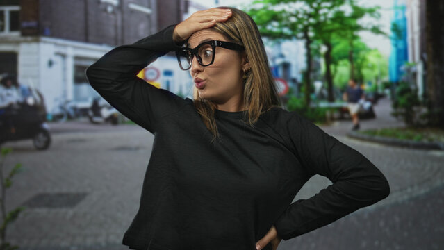 Woman with hand on forehead in a surprised gesture on a busy street wearing dark glasses and casual sweater, facing camera; surprise reaction.
