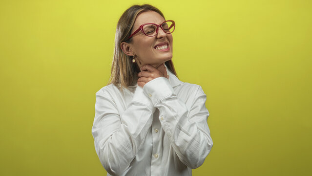 Woman holding throat with hand in yellow studio wearing white shirt and red glasses; sore pain distress.