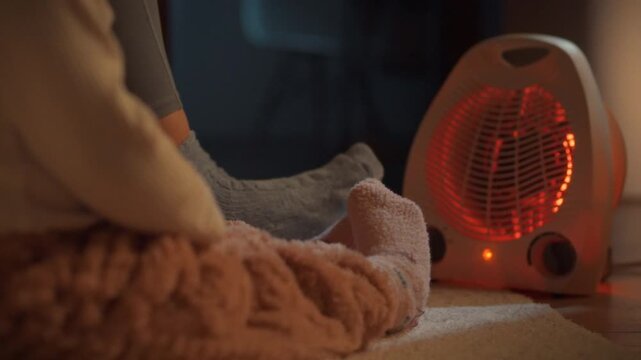 A young mother and child, clad in warm winter socks, snuggle on the sofa, warming their feet beside a bright electric heater with a visible red light on the floor, regulated by a thermostat