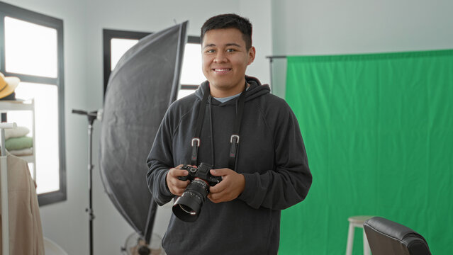 Man holding camera and smiling in studio with green screen, softbox and stool; confidence creativity.