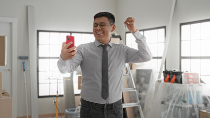 Fototapeta premium Man in dress shirt and tie holding smartphone and keys, right hand raised for selfie while smiling inside a building with ladder and boxes; new home pride.