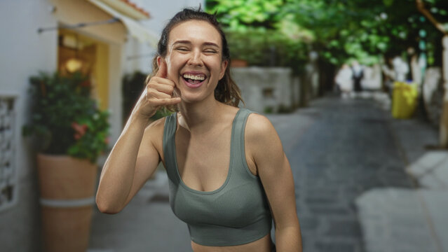 Young hispanic woman smiling and making call sign with her hand in sunlit bustling urban street; playfulness.