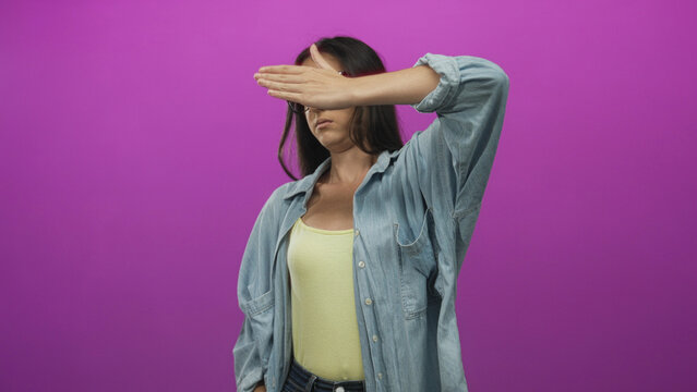 Young hispanic woman covering eyes with hand in studio against purple wall wearing denim shirt and yellow tank; shyness solitude.