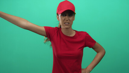 Fototapeta premium Woman courier in red cap and t shirt with puffed cheeks, hands on hips and raised arm in teal green studio; frustration.