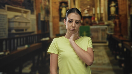 Fototapeta premium Woman with hand on neck and pensive face in church building, yellow shirt and wooden pews visible; uncertainty discomfort.