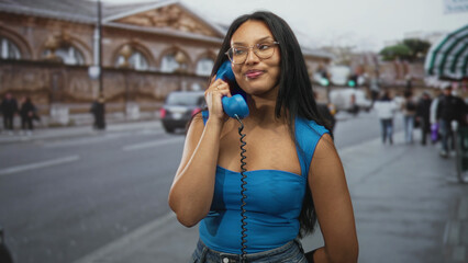 Fototapeta premium Woman holding a blue corded telephone to her ear on a busy street near a historic building; contentment.