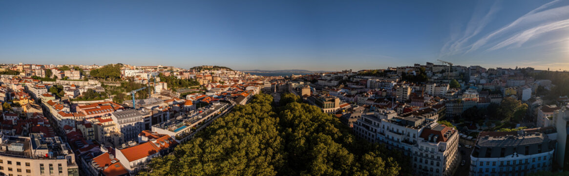 Aerial view of the dense urban landscape featuring red-tiled rooftops, historic buildings, and a lush green park under a clear blue sky during golden hour in Lisbon, Lisbon, Portugal.