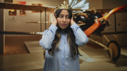Hispanic woman wearing a gold crown covers her ears with both hands in a museum building beside a vintage biplane exhibit  annoyance. © Krakenimages.com