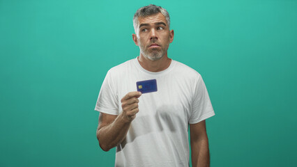 Fototapeta premium Man holds blue creditcard between fingers in a studio teal backdrop, white tshirt visible, hand raised and lips pursed showing uncertainty; financial concern.