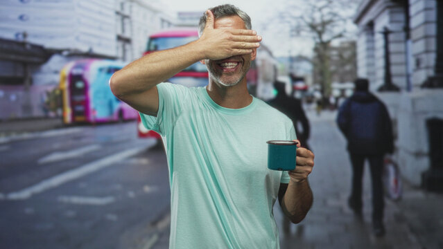 Man with grey hair holding blue mug in hand on city street by a red doubledecker bus, alternately covering his eyes and waving toward camera; playfulness.