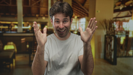 Naklejka premium Man smiling with hands raised and palms open in a hotel building lobby near reception desk and wooden beams; joyful humor candid.