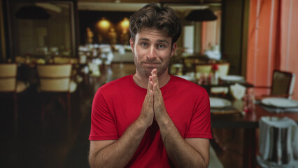 Fototapeta premium Man in red t shirt with hands clasped and grimace seated at a restaurant table inside a building; nervousness.