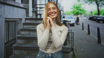 Fototapeta premium Woman clasping hands near chin smiling on stone steps by sidewalk with parked cars on city street; gentle joy.