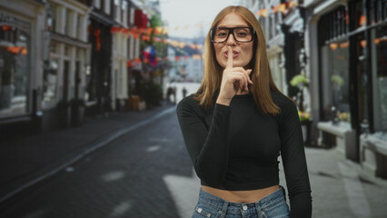 Fototapeta premium Young woman with red hair and glasses places finger to lips for silence on a cobblestone street, wearing black crop top and jeans; playful secrecy.