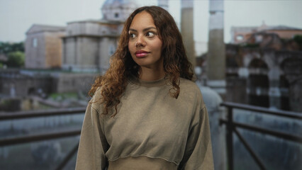 Obraz premium Woman looking sideways with a subtle smile, wearing a loose sweatshirt on a building terrace overlooking roman ruins and columns; serenity.