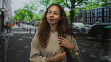 Fototapeta premium Young woman touches hair with both hands on a city street lined with trees and parked cars; confidence wellness.