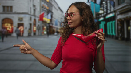 Fototapeta premium Woman holding red high heel shoe points finger to left on street, smiling in red top and glasses; confidence shopping joy.